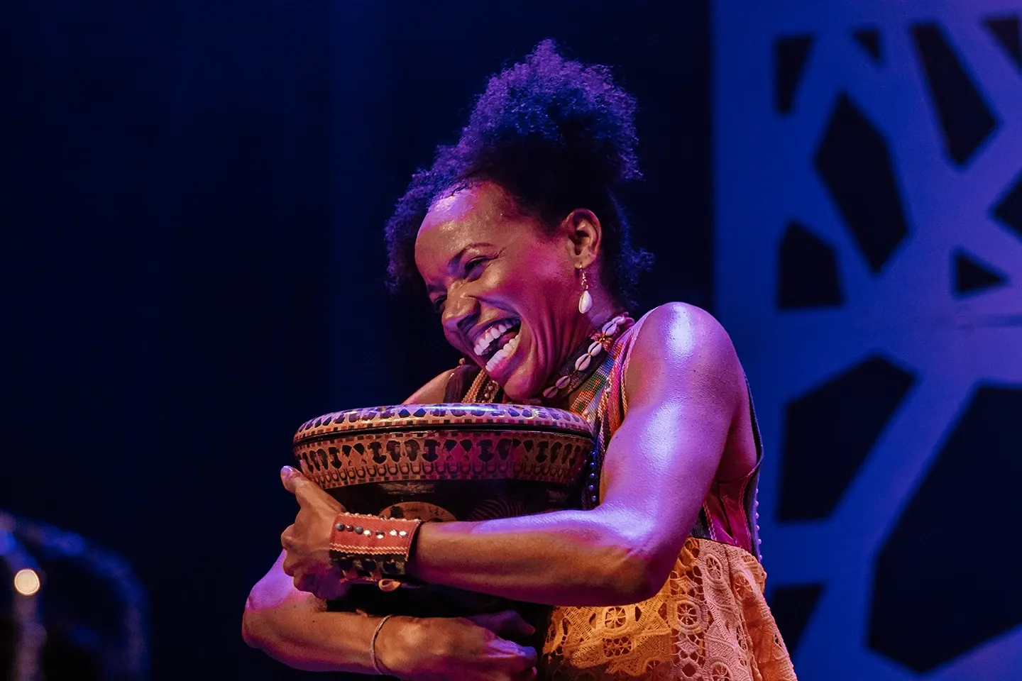 Atriz em espetáculo teatral abraçando tigela artesanal decorada com padrões coloridos, sorrindo amplamente, usando acessórios étnicos e vestido rendado, sob iluminação cênica azul e roxa com cenário recortado ao fundo.