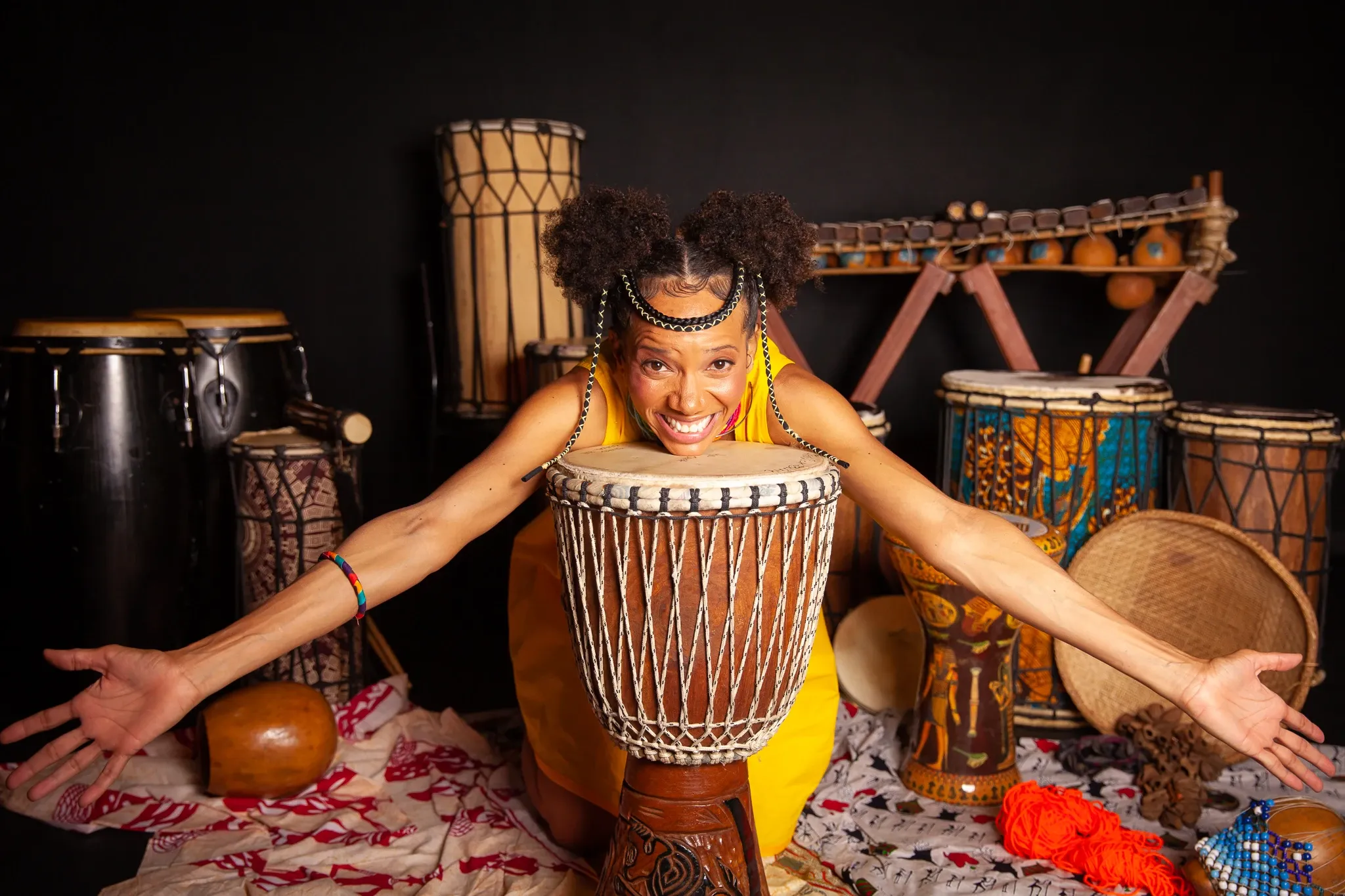 Akili sorridente apoiada sobre djembê com braços abertos, vestindo roupa amarela, rodeada por diversos instrumentos de percussão africanos como conga, balafon e dunduns, sobre tecido estampado em fundo escuro.