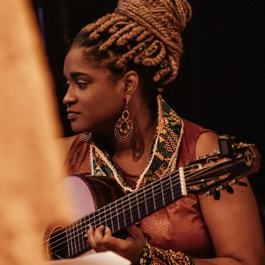 Musicista tocando violão clássico em apresentação ao vivo, com tranças louras presas em coque elaborado, usando vestido bordô com detalhes em tecido africano estampado e brincos dourados, em fundo escuro com iluminação quente.