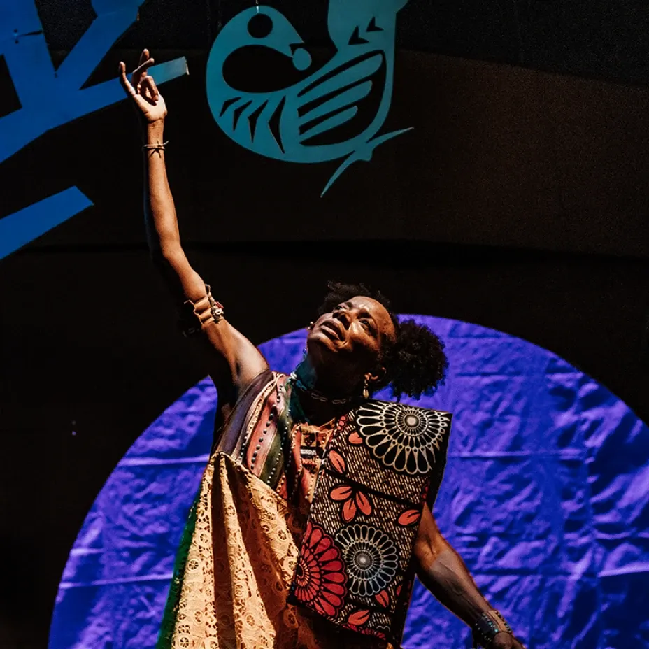 Artista cênica com braço erguido em gesto expressivo durante espetáculo teatral, vestindo trajes africanos estampados coloridos, com cenário iluminado em azul e roxo e figura de pássaro estilizado ao fundo.