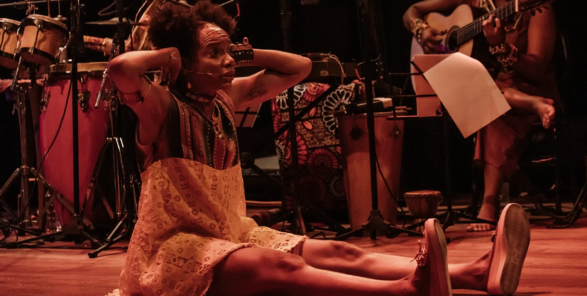 Atriz sentada no chão do palco com expressão intensa e mãos na cabeça, vestindo trajes étnicos coloridos, rodeada por instrumentos de percussão e músicos ao fundo, sob iluminação cênica quente avermelhada em espetáculo teatral.