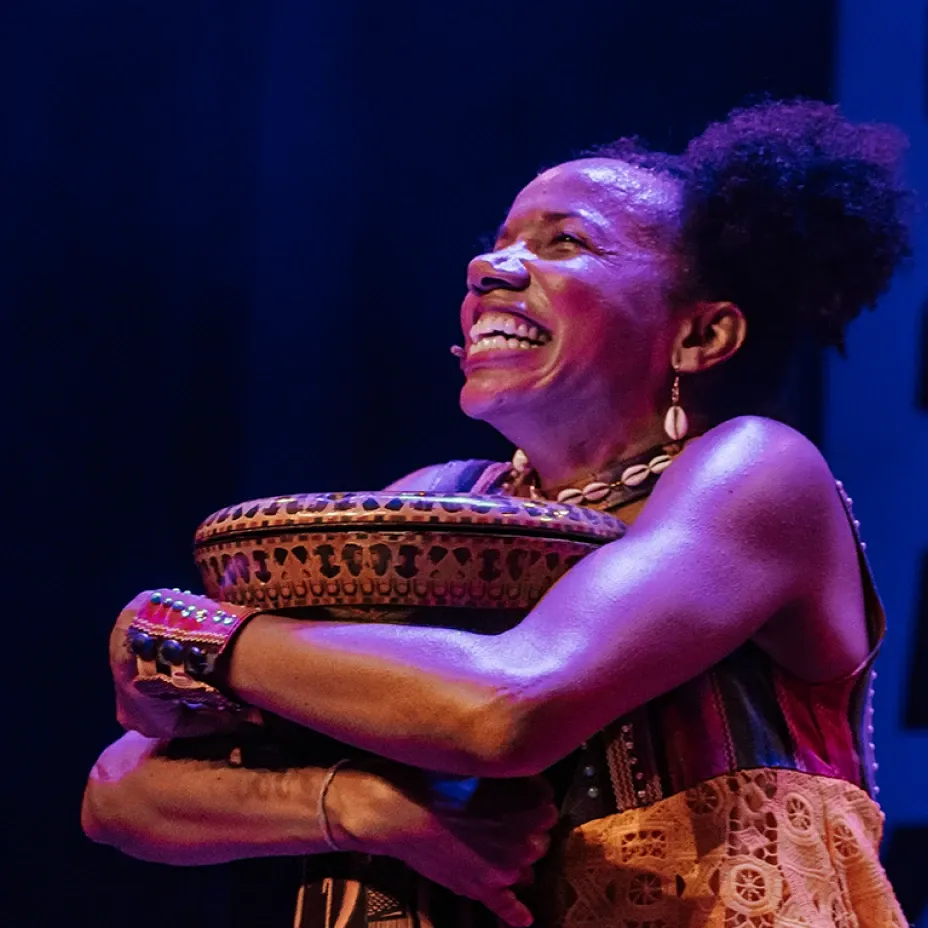 Artista teatral sorrindo com olhar voltado para o alto enquanto abraça tigela artesanal decorada, usando colar de búzios e pulseiras étnicas, sob iluminação cênica azul e roxa em fundo escuro.