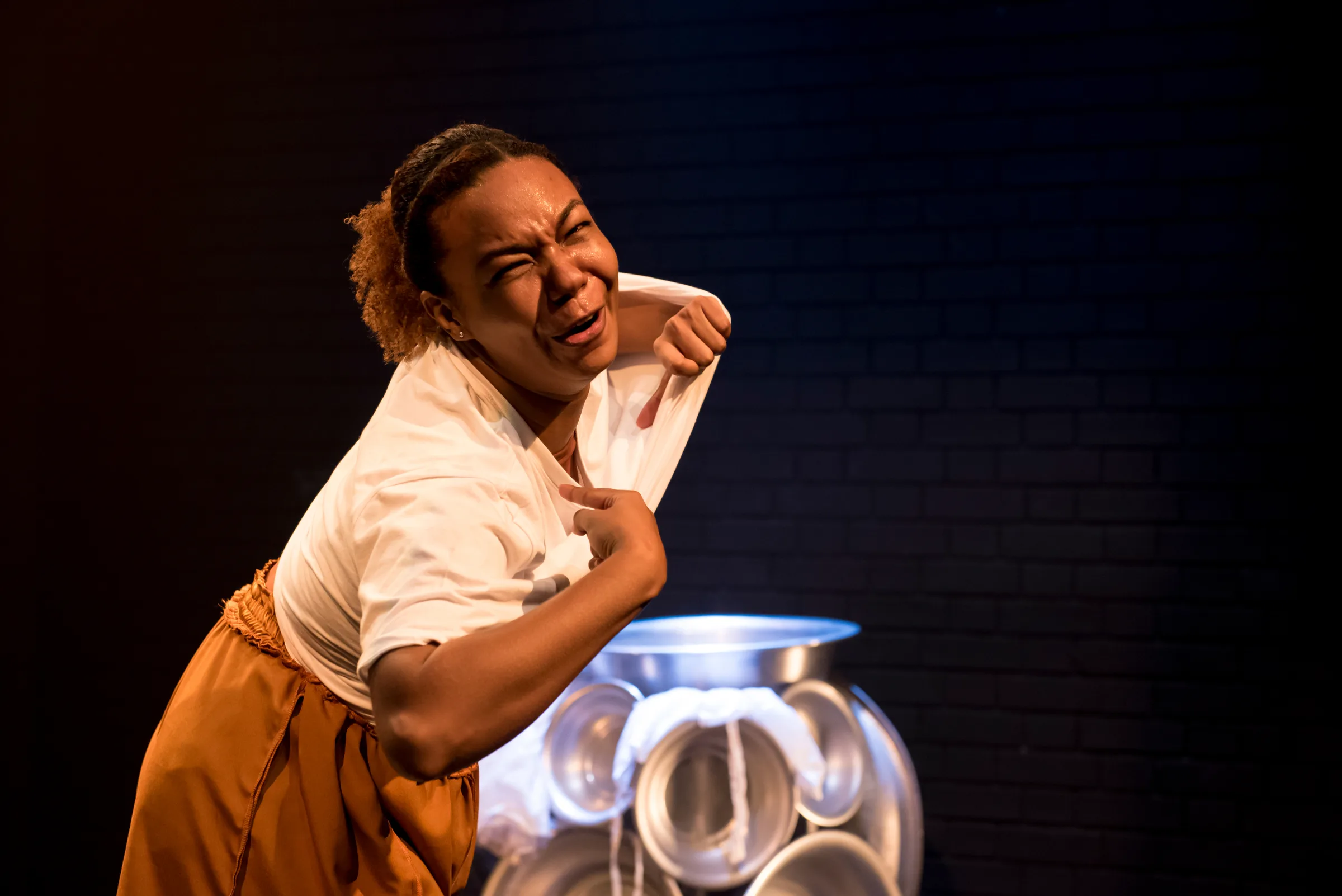 Atriz em cena teatral expressando emoção intensa, vestindo camiseta branca e saia caramelo, com escultura metálica iluminada ao fundo em palco com parede de tijolos escura.