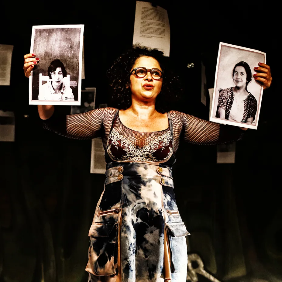 Uma mulher em cena teatral segura duas fotografias em preto e branco, uma em cada mão, enquanto está diante de um fundo escuro com papéis pendurados. Ela usa óculos e um figurino artístico, e parece estar apresentando ou comparando as imagens ao público.
