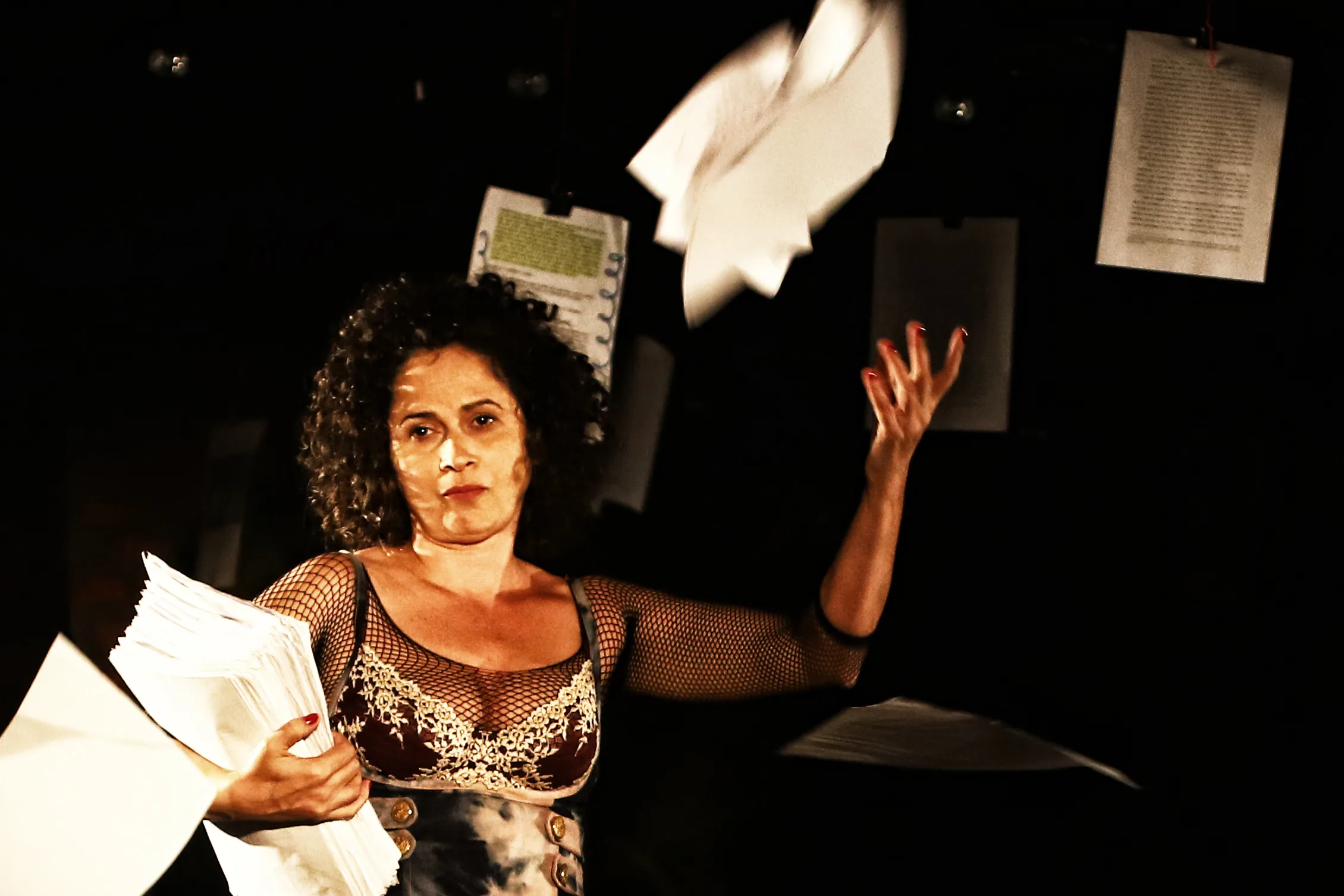 Uma performer em cena teatral segura um monte de papéis enquanto lança outros ao ar. Ela veste figurino artístico com mangas em rede e está diante de um fundo escuro com folhas penduradas, criando uma atmosfera dramática.