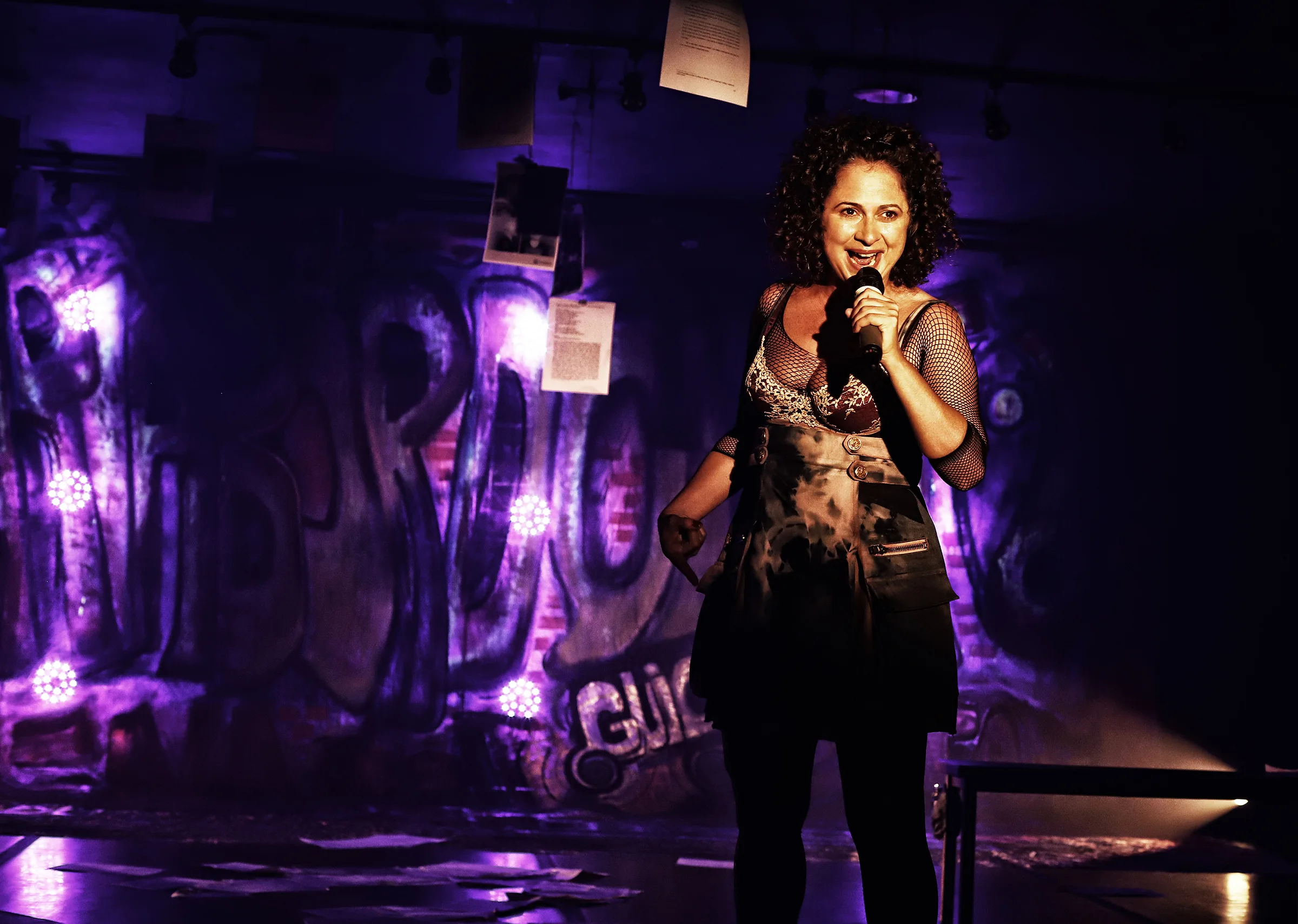 Uma performer em pé no palco segura um microfone e sorri enquanto se apresenta. Ela veste figurino artístico com mangas em rede, sob iluminação roxa, com papéis pendurados ao fundo criando um cenário teatral.
