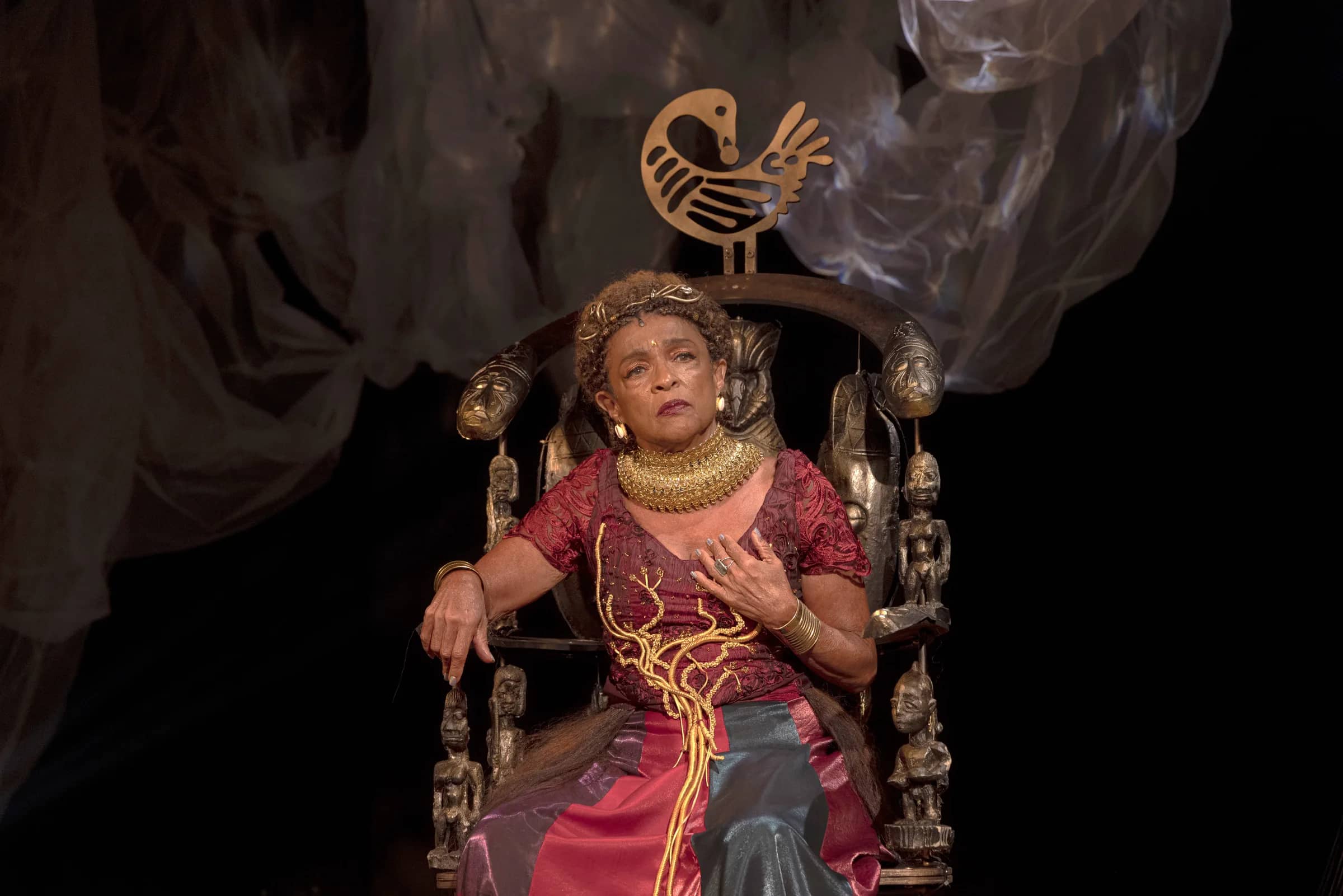 Atriz idosa sentada em um trono decorado com esculturas, vestindo traje vermelho e dourado com colar volumoso, em um palco escuro com tecidos translúcidos ao fundo, expressão séria e mão sobre o peito.
