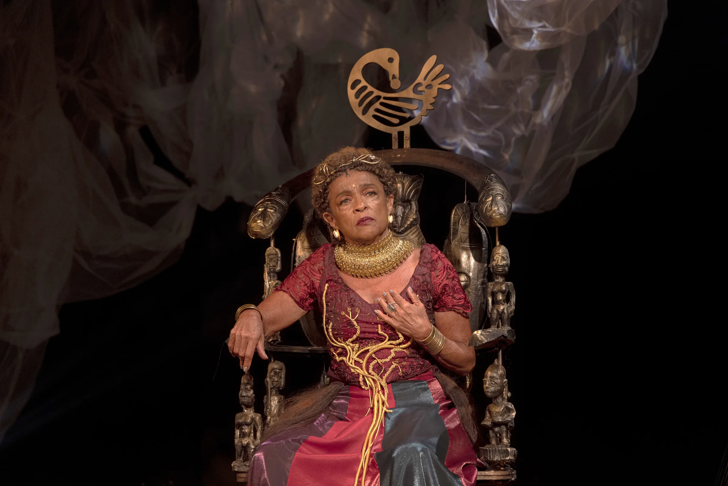 Atriz idosa sentada em um trono decorado com esculturas, vestindo traje vermelho e dourado com colar volumoso, em um palco escuro com tecidos translúcidos ao fundo, expressão séria e mão sobre o peito.